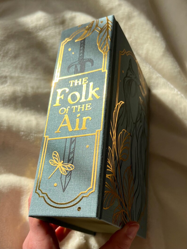 Puede incluir: Un libro de tapa dura azul verdoso con letras e ilustraciones doradas. El t&iacute;tulo es "The Folk of the Air".