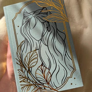 Puede incluir: Un diario azul claro con un dise&ntilde;o de l&aacute;mina dorada de una mujer con cabello largo y hojas. La mujer tiene cuernos y est&aacute; enmarcada en un &oacute;valo dorado.