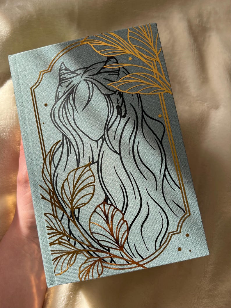 Puede incluir: Un diario azul claro con un dise&ntilde;o de l&aacute;mina dorada de una mujer con cabello largo y hojas. La mujer tiene cuernos y est&aacute; enmarcada en un &oacute;valo dorado.