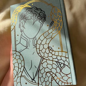 Puede incluir: Un libro de tapa dura azul turquesa con un dise&ntilde;o en l&aacute;mina dorada de una persona con una serpiente enroscada a su alrededor. La serpiente tiene un patr&oacute;n de escamas detallado.