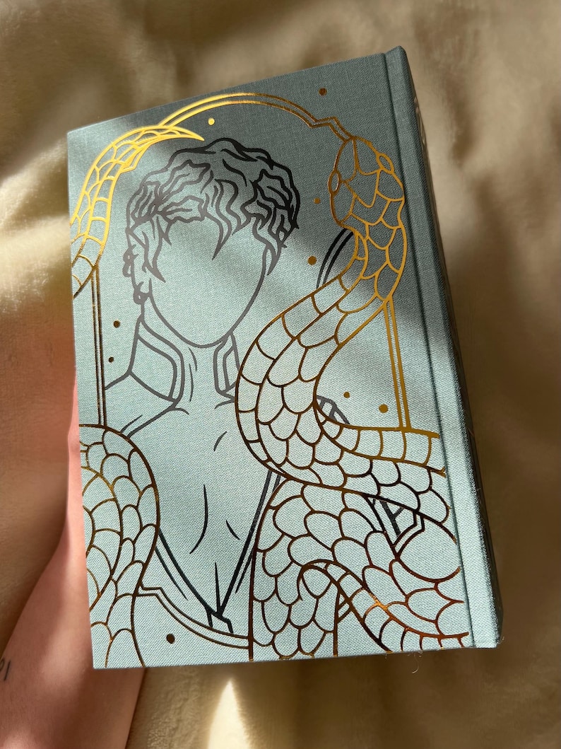 Puede incluir: Un libro de tapa dura azul turquesa con un dise&ntilde;o en l&aacute;mina dorada de una persona con una serpiente enroscada a su alrededor. La serpiente tiene un patr&oacute;n de escamas detallado.