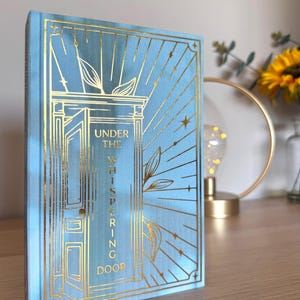 Könnte beinhalten: Ein Hardcover-Buch mit dem Titel "Under the Whispering Door" mit hellblauem Einband und goldenen Folienakzenten. Das Cover zeigt ein verziertes Türdesign mit Strahlenlinien und botanischen Elementen. Das Buch liegt auf einer Holzoberfläche.