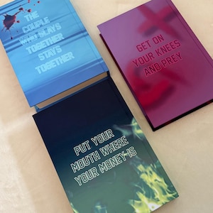 May include: Three hardcover books with distinct covers. The blue book features a blood splatter and the text THE COUPLE WHO SLAYS TOGETHER STAYS TOGETHER. The purple book displays the text GET ON YOUR KNEES AND PREY. The dark book shows green flames and the text PUT YOUR MOUTH WHERE YOUR MONEY IS.