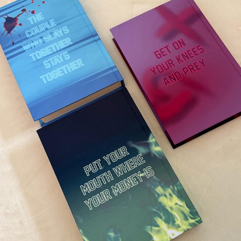 May include: Three hardcover books with distinct covers. The blue book features a blood splatter and the text THE COUPLE WHO SLAYS TOGETHER STAYS TOGETHER. The purple book displays the text GET ON YOUR KNEES AND PREY. The dark book shows green flames and the text PUT YOUR MOUTH WHERE YOUR MONEY IS.