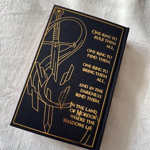 May include: A black book cover with gold lettering and a gold illustration of a sword. The text reads "One Ring to Rule Them All, One Ring to Find Them, One Ring to Bring Them All, And in the Darkness Bind Them, In the Land of Mordor Where the Shadows Lie."