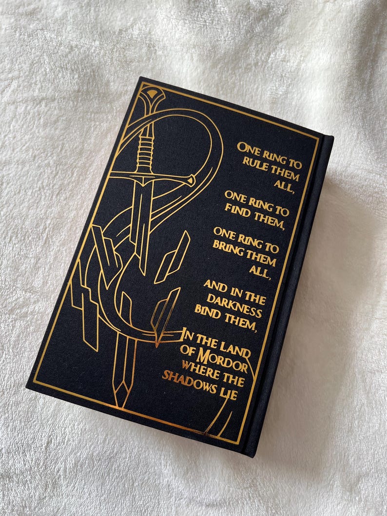 May include: A black book cover with gold lettering and a gold illustration of a sword. The text reads "One Ring to Rule Them All, One Ring to Find Them, One Ring to Bring Them All, And in the Darkness Bind Them, In the Land of Mordor Where the Shadows Lie."