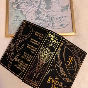 May include: A black hardcover book set of "The Lord of the Rings" by J.R.R. Tolkien. The book covers feature gold lettering and illustrations of a sword, a flower, and a ring.