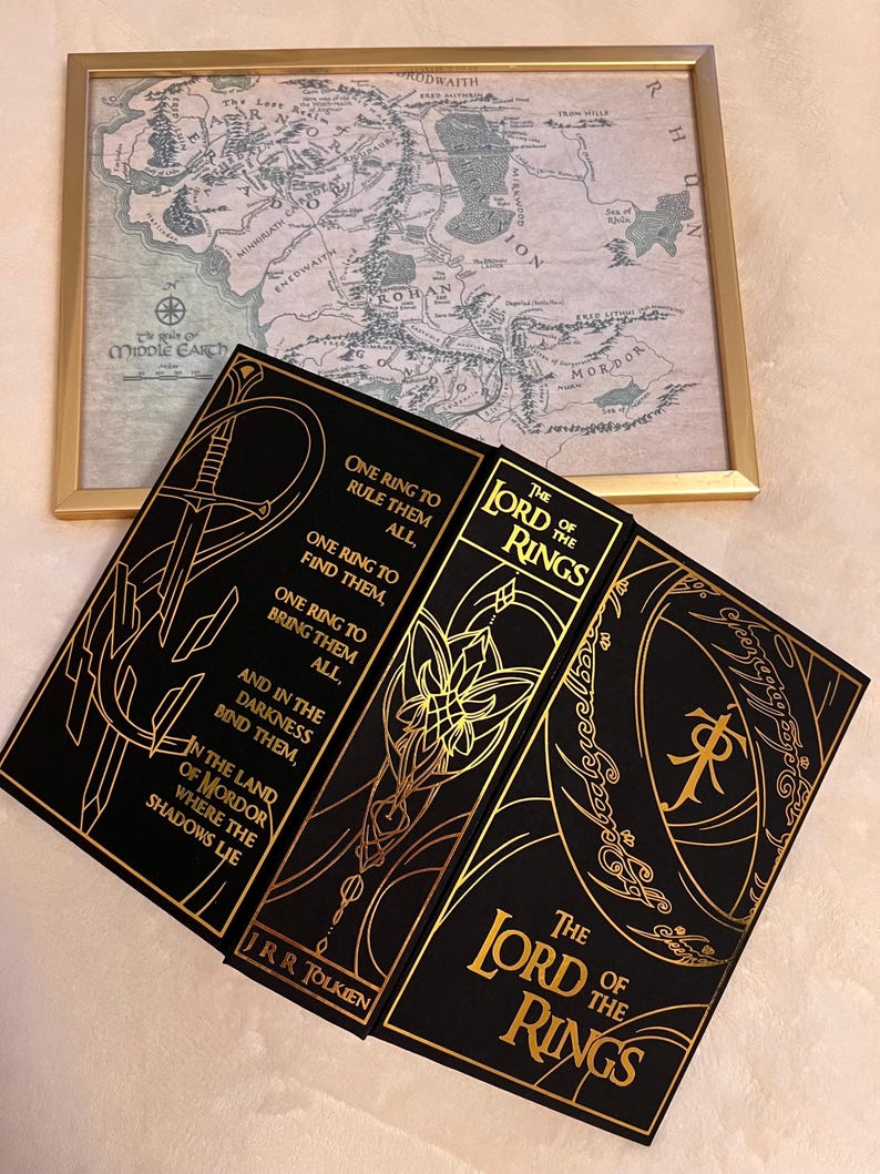 May include: A black hardcover book set of "The Lord of the Rings" by J.R.R. Tolkien. The book covers feature gold lettering and illustrations of a sword, a flower, and a ring.