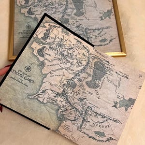 May include: A vintage-style map of Middle-earth from The Lord of the Rings, featuring the text "The Realm of Middle-earth" and "The Lost Realm of Numenor". The map is printed on a cream-colored paper with brown and black ink.