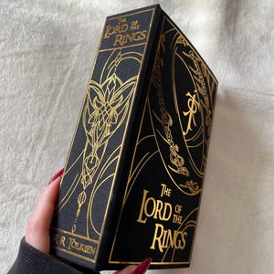 May include: A black hardcover book with gold lettering and illustrations. The title is "The Lord of the Rings" by J.R.R. Tolkien.