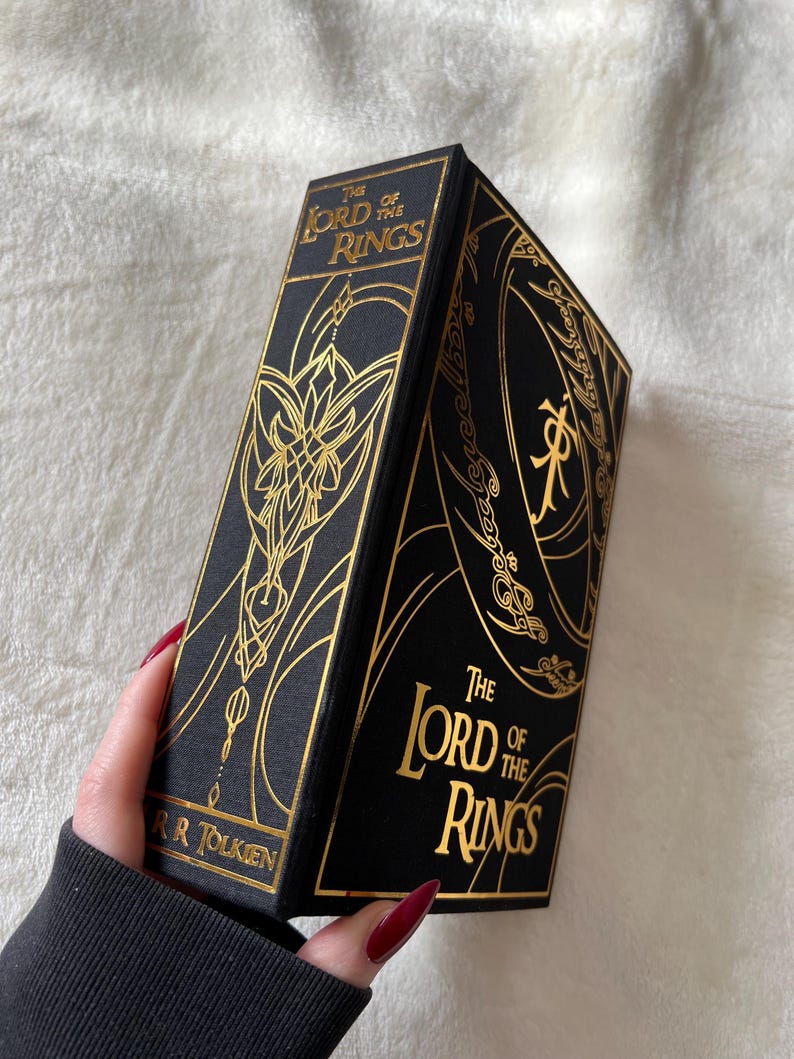 May include: A black hardcover book with gold lettering and illustrations. The title is "The Lord of the Rings" by J.R.R. Tolkien.