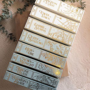 May include: A collection of eight books with decorative covers. The books are in varying shades of blue and white, with gold lettering and illustrations. Titles include "Kingdom of Ash," "Tower of Dawn," and "Empire of Storms."
