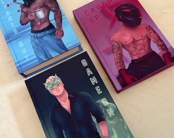 May include: Three hardcover books with illustrated covers. The blue book features a masked figure holding a mug with the text LIGHTS OUT. The magenta book shows a helmeted figure with the text CAUGHT UP. The black book shows a masked figure with GAME ON.