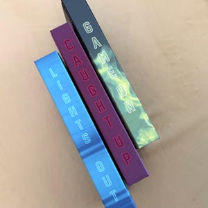 May include: Three rectangular boxes arranged diagonally. The blue box is labeled LIGHTS OUT, the purple box is labeled CAUGHT UP, and the dark green box is labeled GAME ON. Each box features white outlined lettering printed vertically on the surface.