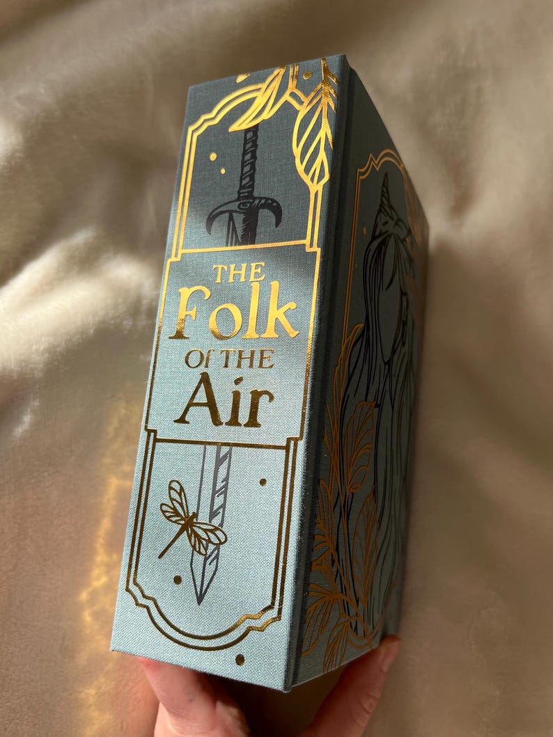 Puede incluir: Un libro de tapa dura azul con letras doradas e ilustraciones. El t&iacute;tulo es "The Folk of the Air".