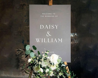 Puede incluir: Un letrero de boda gris con el texto "WELCOME TO THE WEDDING OF DAISY & WILLIAM" en blanco. El letrero está apoyado en un caballete de madera y decorado con flores blancas y vegetación.