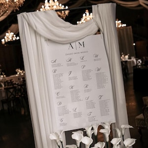 Könnte beinhalten: Ein weißes Hochzeits-Sitzplan mit schwarzer Schrift, präsentiert auf einem weißen drapierten Hintergrund. Die Tafel ist mit weißen Blumen am unteren Rand verziert. Der Text oben lautet "AM" und "ZNAJDŹ SWOJE MIEJSCE".