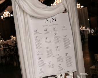 May include: A white wedding seating chart with black lettering, displayed on a white draped backdrop. The chart is decorated with white flowers at the base. The text at the top reads "AM" and "ZNAJDŹ SWOJE MIEJSCE".