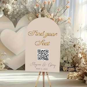 以下が含まれることがあります： 「Find your Seat」と書かれた金色の文字が入ったベージュ色のウェディングサイン。その下にQRコードと「Megan & Greg」の名前、日付「NOV 5, 2025」が記載されています。サインは金のイーゼルに立てられています。