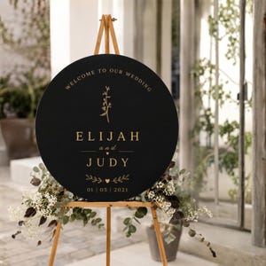 Op de afbeelding: Een zwart, rond trouwbord op een houten ezel. Het bord heeft gouden letters met bovenaan "WELCOME TO OUR WEDDING" en in het midden de namen "ELIJAH and JUDY". De datum "01 | 05 | 2021" staat onderaan.