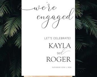 May include: A white sign on a wooden easel with the words "we're engaged" in elegant script. Below, it reads "LET'S CELEBRATE!" followed by "KAYLA and ROGER" and the date "SATURDAY JUNE 1, 2024". The background is lush green foliage.