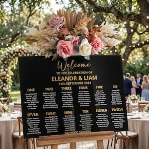 May include: A black wedding seating plan with gold lettering, listing guest names and table assignments. The sign reads "Welcome to the Celebration of Eleanor & Liam, 14th September 2024." The sign is decorated with a floral arrangement and stands on a wooden easel.