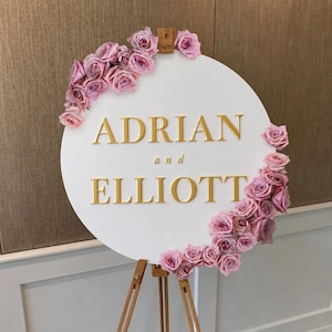 Op de afbeelding: Een rond wit bord met de gouden tekst ADRIAN en ELLIOTT, met het woord and in kleinere gouden letters ertussen. Het bord is versierd met trossen lichtpaarse rozen langs de linkerbovenrand en de rechteronderrand.