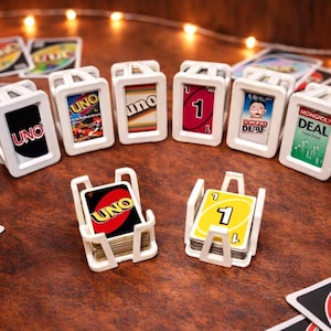 Może przedstawiać: Białe uchwyty na karty z kartami Uno i Monopoly Deal. Karty mają odważne kolory i logo Uno. Uchwyty są ułożone na ciemnej drewnianej powierzchni, z rozrzuconymi kartami w tle.