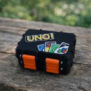 Puede incluir: Estuche rectangular negro con la palabra "UNO!" en amarillo y blanco. El estuche tiene cierres naranjas y contiene varias cartas de UNO con varios colores y símbolos. El estuche está sobre una superficie de madera.