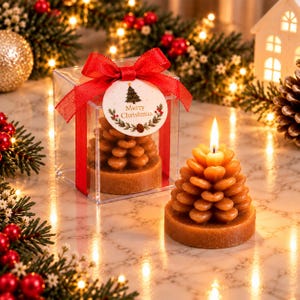 Puede incluir: Una vela marrón con forma de piña se encuentra junto a una caja de regalo de plástico transparente que contiene una vela idéntica. La caja está atada con una cinta roja y tiene una etiqueta redonda con un árbol y Merry Christmas.