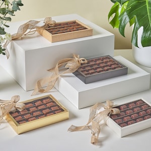 Peut inclure: Quatre boîtes de chocolats assortis, chacune attachée d'un ruban brun. Les boîtes sont disposées sur trois plateformes blanches. Les boîtes sont faites de différents matériaux, dont du carton, du bois et du métal. Les chocolats sont visibles à travers les fenêtres en plastique transparent des boîtes.