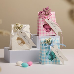 Puede incluir: Tres pequeñas cajas de regalo con ventanas en forma de corazón y etiquetas de "Gracias". Las cajas están decoradas con patrones de cuadros vichy en rosa, azul y blanco. Cada caja tiene un pequeño ramo de flores blancas y una cinta.