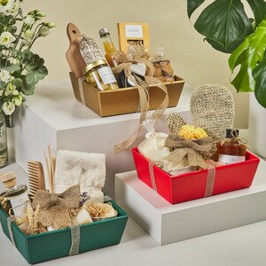 May include: Three gift baskets filled with various bath and body products. The baskets are green, red, and gold. The green basket contains a bottle of essential oil, a comb, a towel, and other items. The red basket contains a bottle of body wash, a loofah, and other items. The gold basket contains a bottle of wine, a bottle of olive oil, a wooden cutting board, and other items.