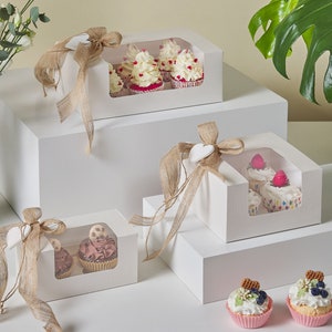 Pode incluir: Quatro caixas de cartão branco com janelas transparentes que mostram cupcakes decorados. As caixas são amarradas com barbante e têm uma etiqueta em forma de coração. As caixas estão empilhadas umas sobre as outras em uma superfície branca. Dois cupcakes adicionais estão na superfície em frente às caixas.