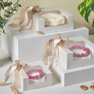 Op de afbeelding: Drie witte kartonnen cake dozen met doorzichtige plastic ramen, elk versierd met een jute lint en een wit hart. De dozen zijn op elkaar gestapeld op een wit oppervlak. De bovenste doos bevat een cake met witte frosting, de middelste doos bevat een cake met rode en blauwe bessen, en de onderste doos bevat een cake met een vergelijkbaar ontwerp als de middelste doos.
