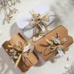 May include: Three gift boxes wrapped in brown and white corrugated cardboard with a rustic twine bow and dried flowers. The boxes have small tags that say "Thank you".