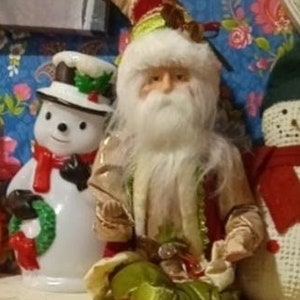 May include: A festive Christmas scene with a Santa Claus doll wearing a green and red outfit, a snowman doll with a red scarf and a wreath, and a white ghost doll. The dolls are sitting on a white shelf with a string of lights and a pumpkin.