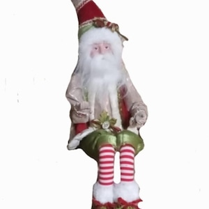 Könnte beinhalten: Eine dekorative Weihnachtsmann-Figur mit einer langen, spitzen beigen Mütze, die mit einem weißen Bommel versehen ist. Die Figur hat einen weißen Bart, einen rot-beigen Mantel, eine grüne Hose und rot-weiß gestreifte Beine mit grünen Paillettenschuhen.