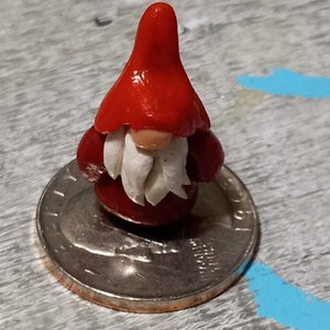 May include: A small red gnome figurine with a white beard and a red hat. The gnome is sitting on a US quarter.