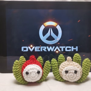 May include: Two crocheted plush toys, one red and one white, with green leafy accents. The toys are in front of a tablet screen displaying the Overwatch logo.