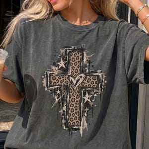 May include: A charcoal grey short-sleeve t-shirt featuring a large central graphic of a cross with a leopard print pattern, a heart shape in the centre, and several star accents. The shirt has a faded, washed-out appearance and a crew neckline.