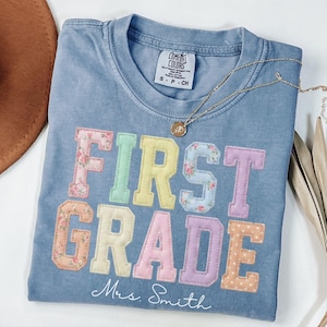 May include: A blue jean colored t-shirt featuring the text FIRST GRADE in large, multi-colored pastel block letters with floral and polka dot patterns. Below the text, the name Mrs. Smith is printed in white cursive script. A gold necklace rests on the collar.