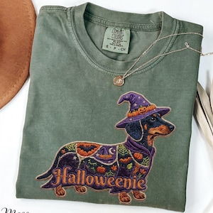 May include: A moss green short-sleeve t-shirt featuring a graphic of a dachshund wearing a purple witch hat and a patterned cape with pumpkins, bats, and spiderwebs. The text "Halloweenie" is printed in orange below the dog illustration.