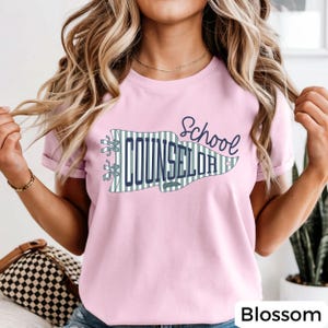 May include: A light pink short-sleeve t-shirt featuring a graphic of a pennant flag with green and white vertical stripes. The text "School COUNSELOR" is printed in dark blue, with a small star icon near the right edge of the pennant.