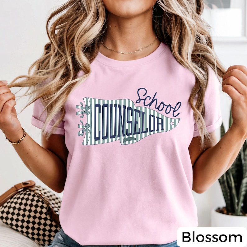May include: A light pink short-sleeve t-shirt featuring a graphic of a pennant flag with green and white vertical stripes. The text "School COUNSELOR" is printed in dark blue, with a small star icon near the right edge of the pennant.