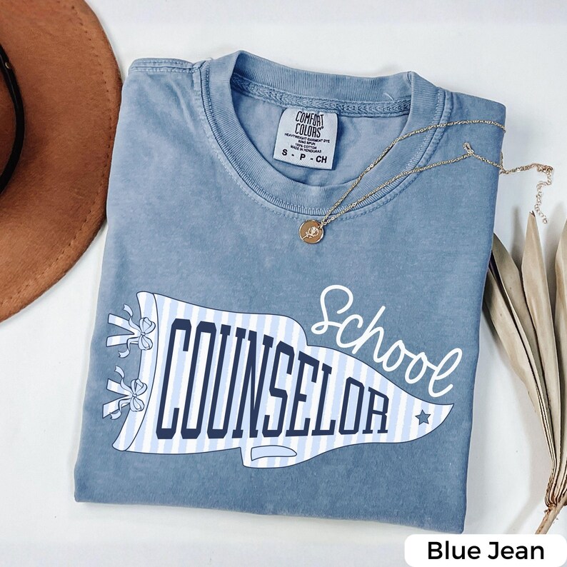 May include: A blue jean coloured t-shirt with a Comfort Colors label at the neck. The front features a white and light blue striped pennant graphic with the text School COUNSELOR in dark blue, accented by two small bows and a star.