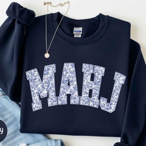 May include: A navy blue crewneck sweatshirt featuring the letters MAHJ in a large, arched collegiate font. The letters are filled with a white and blue floral pattern. A thin gold necklace with a small circular pendant rests above the neckline.