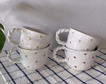 May include: Four white ceramic mugs with floral patterns. The mugs have a twisted rope handle and are stacked in pairs. The mugs are on a white and blue checkered tablecloth.