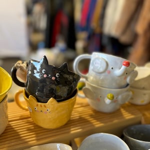 May include: A selection of ceramic mugs featuring animal designs. The mugs include a black cat, a yellow cat, and an elephant. The mugs are stacked on a wooden shelf, with other mugs visible in the background.