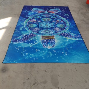 Op de afbeelding: Een blauw en groen mandala-achtig zeeschildpadontwerp op een blauwe achtergrond met witte bubbels. De schildpad heeft een gouden ketting en ingewikkelde details.